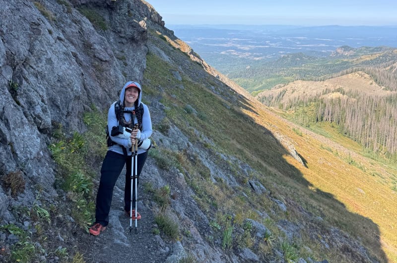 Hiker standing on a narrow mountain trail with trekking poles, steep rocky slope and long-distance views behind.