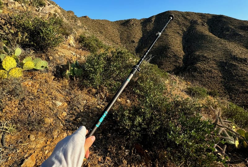 Hiker using a trekking pole on a sunny desert trail surrounded by dry brush and cactus.