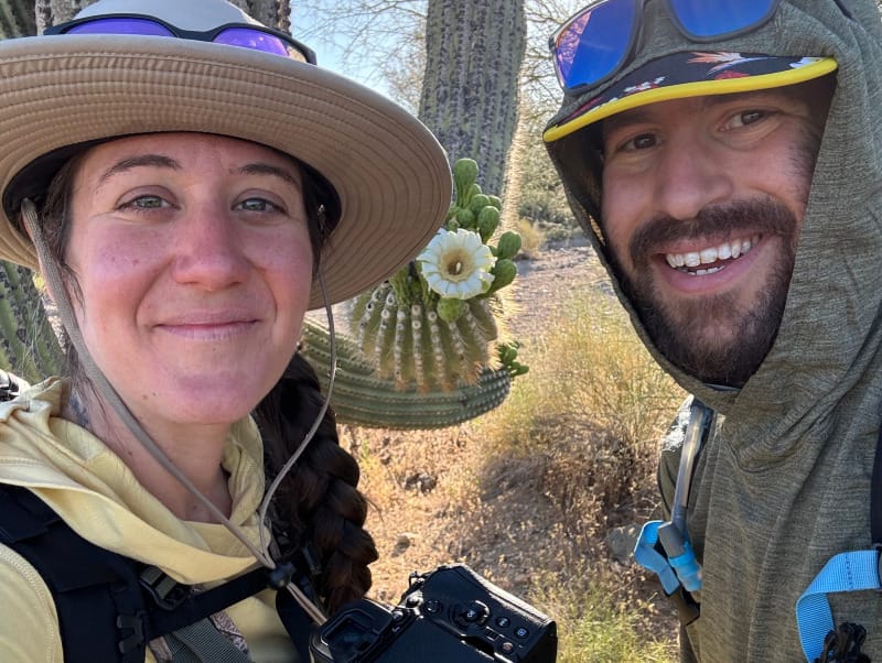 Two hikers wearing sun-protective hats and lightweight gear standing near blooming saguaro cacti in morning light.
