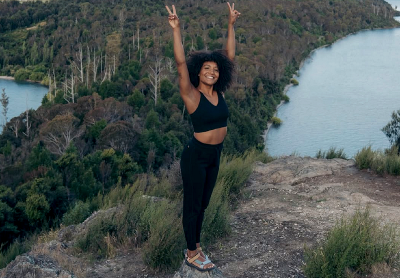  Woman standing on a cliffside wearing Branwyn Essential Compression Leggings with arms raised in a peace sign, overlooking a scenic lake and mountain view.