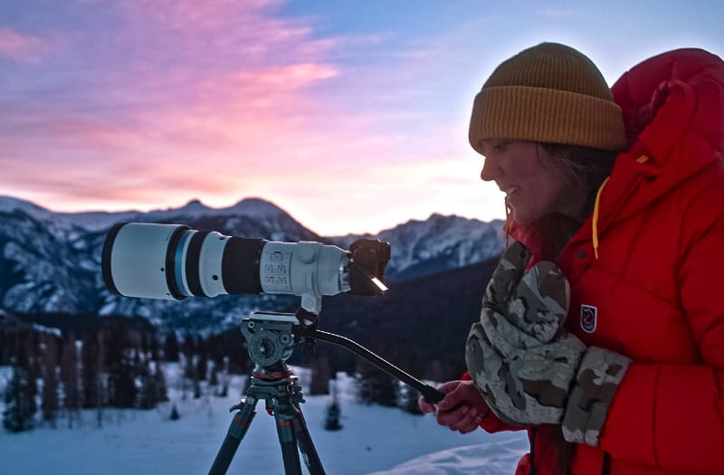 Photographer using a tripod at sunset in snowy mountains while wearing KUIU Super Down Pro Glomitts, demonstrating their warmth and dexterity in winter conditions.