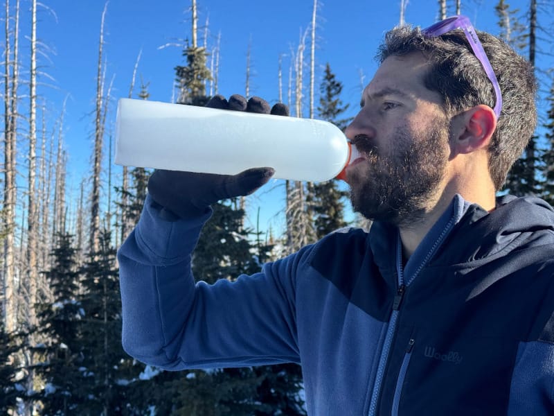 Hiker drinking from a CNOC Outdoors ThruBottle in snowy winter conditions with evergreen trees in the background.