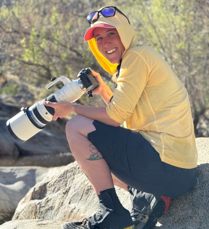 Hiker wearing a yellow sun hoodie in intense desert sun while photographing wildlife on rocky terrain, demonstrating sun protection in hot conditions.
