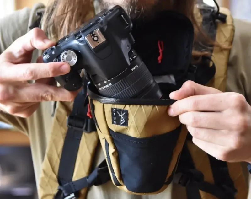 Hiker placing a camera into a Chicken Tramper Ultralight Gear chest-mounted camera pouch designed for backpacking and trail photography.