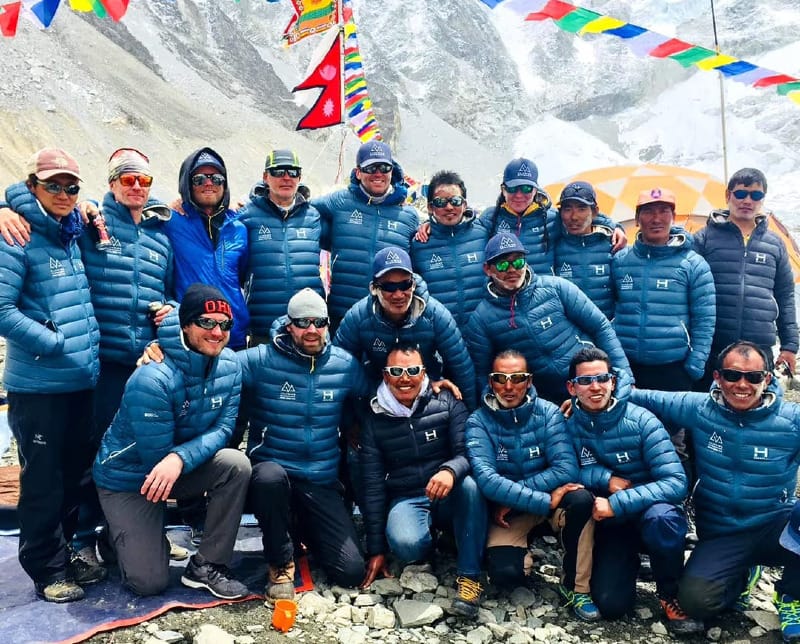 HIMALI team and Himalayan climbers wearing insulated jackets at high altitude with prayer flags and snow-covered peaks in the background