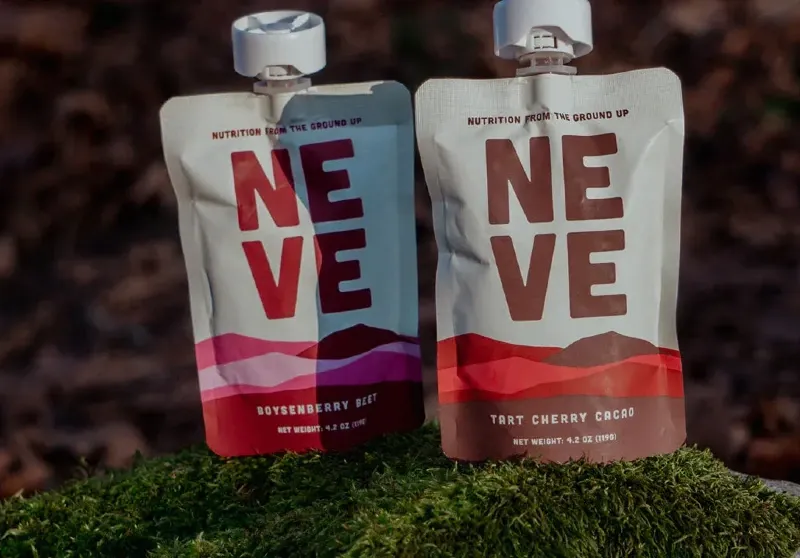 Two Neve Foods smoothie pouches sitting on a moss-covered rock, featuring Boysenberry Beet and Tart Cherry Cacao flavors.