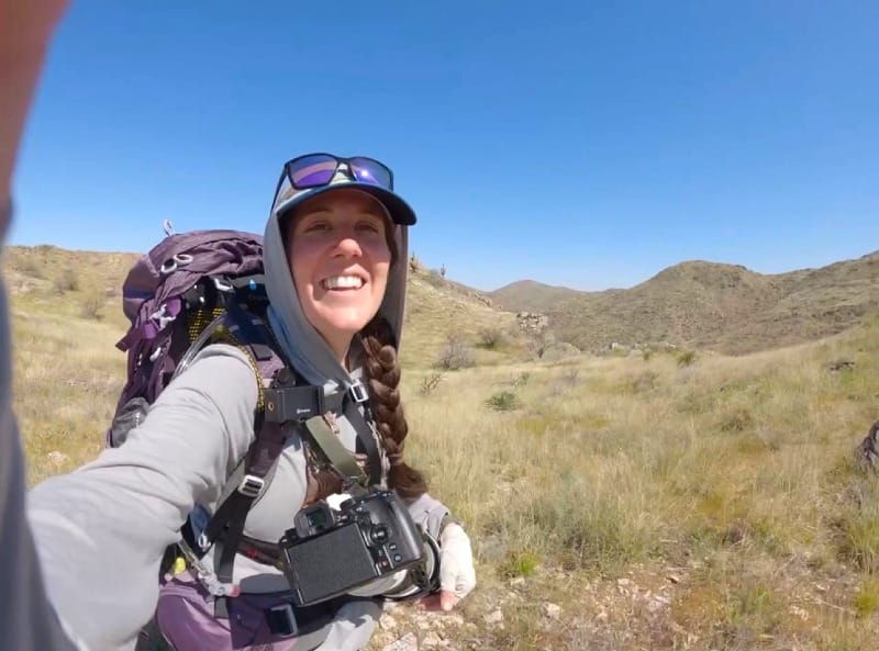 Backpacker wearing a gray sun hoodie while hiking through open desert terrain with a camera and backpack under bright midday sun.