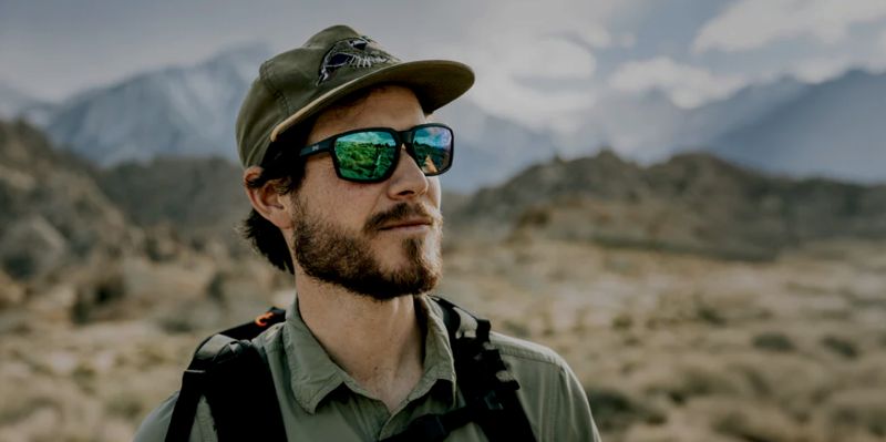 Man wearing green mirrored Wiley X sunglasses and backpack while hiking in a mountainous desert landscape.