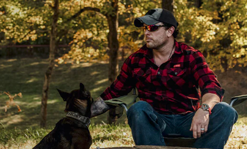 Man wearing red mirrored Wiley X sunglasses and plaid shirt sitting outdoors in a wooded setting with his dog.