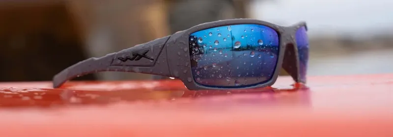 Close-up of Wiley X sunglasses with water droplets on blue mirrored lenses resting on a red surface, highlighting durable frame and wrap design.