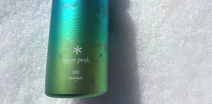 Snow Peak Aurora Titanium Water Bottle in blue-green gradient color laying flat on snow