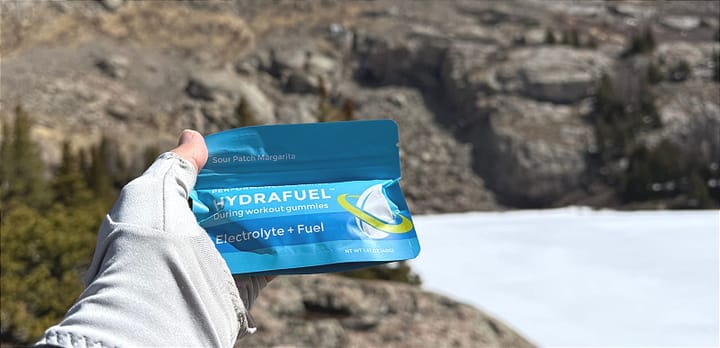  HydraFuel Hydration Gummies package held up in front of a frozen alpine lake and mountain landscape by a gloved hiker.