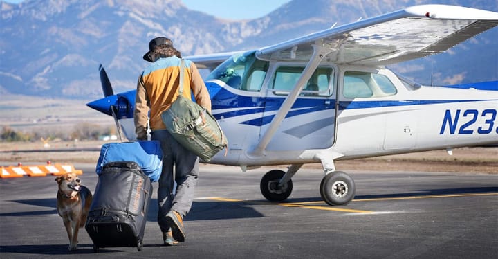  Person walking toward a small plane with Mystery Ranch travel bags and a dog, set against a backdrop of mountains, highlighting durable outdoor gear in use.