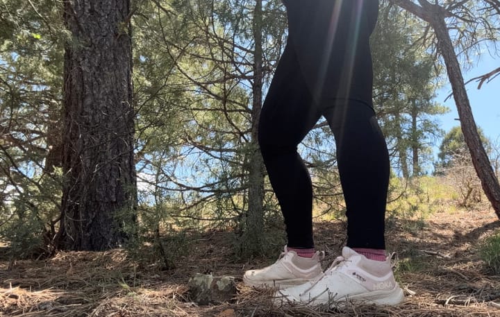  Person standing in forest wearing Ibex Enduro Tights and white trail runners, framed by sunlight and pine trees.