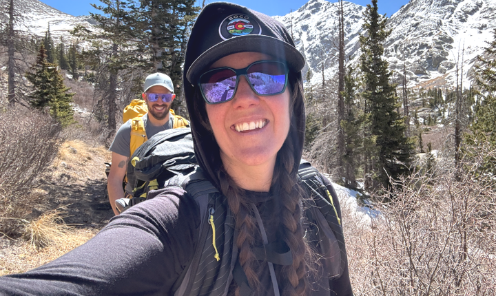  Smiling hiker wearing the black Ridge Merino Solstice Hoodie on a sunny alpine trail with snowy peaks in the background.