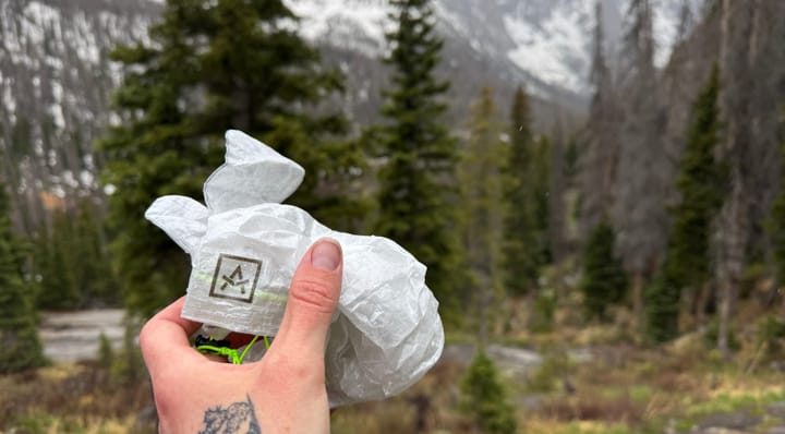 Hand holding Adotec Gear Rain Mitt Stuff Sacks in front of snow-dusted mountains and evergreen trees