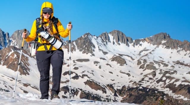  Hiker wearing yellow Merino hoodie trekking through snow with poles, surrounded by dramatic San Juan peaks in the background.