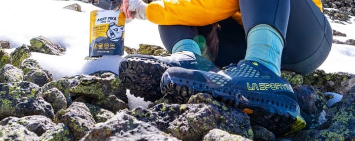  Hiker wearing La Sportiva Spire GTX boots on snowy rocks while preparing Angry Pika dehydrated food in bright sunlight.