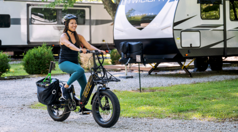  Woman riding a Buzz Centris folding eBike near a campground with RVs in the background, showing off its comfort and utility for RV travel and casual riding.