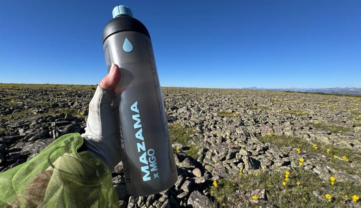  Person holding a Mazama M!GO water bottle in the alpine with rocky terrain and wildflowers in the background.