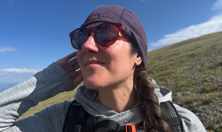 Close-up of a woman hiking with FKT High Lonesome sunglasses and a purple beanie, smiling under blue sky.