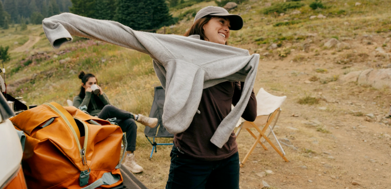 Woman putting on the PAKA Women’s Hoodie at a campsite, with hiking gear and chairs in the background.