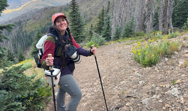 Woman hiking in the mountains wearing KUIU Tiburon Pants, carrying trekking poles and a camera, with rugged alpine terrain in the background.