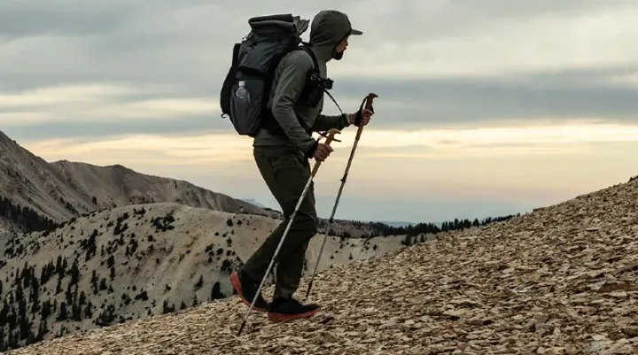 Hiker wearing the Outdoor Vitals Pursuit Hybrid Fleece while climbing a rocky mountain slope at sunrise, carrying a large backpack and trekking poles.