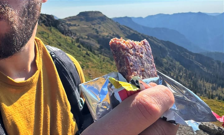 Hiker eating a Skratch Labs Energy Bar on the trail with mountain ridges in the background.