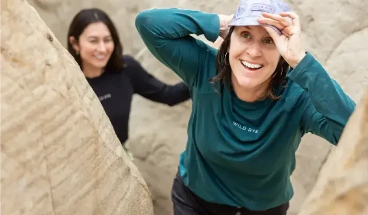  Woman smiling while hiking through a sandstone slot canyon wearing a green Wild Rye long-sleeve and black shorts.