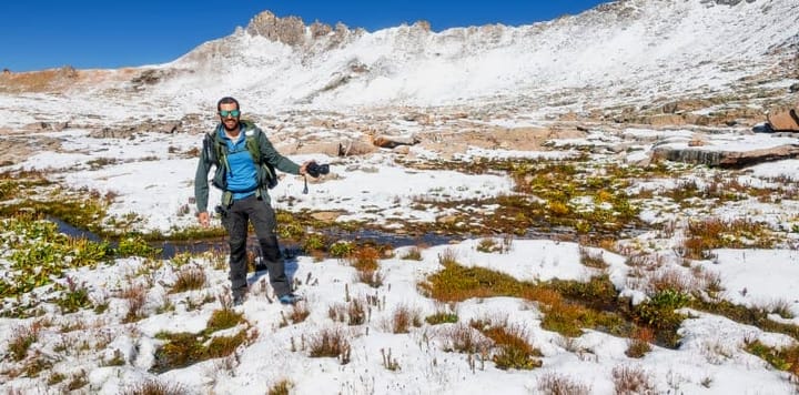 Hiker in layered cold-weather hiking gear standing in a snow-dusted alpine meadow with rugged peaks in the background, autumn mountain adventure.