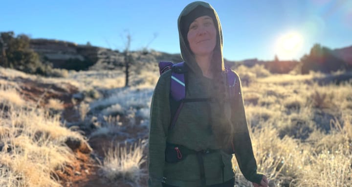 Woman wearing a green wool hoodie and backpack while holding a camera on a frosty desert trail at sunrise.