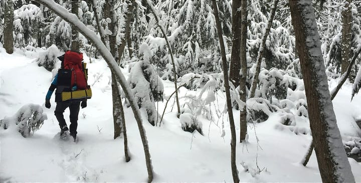 Backpacker with red pack hiking through snowy forest trail — how to hike in cold weather 2025 guide.