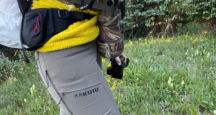 Hiker wearing gray KUIU Attack Pants while carrying camera gear and a backpack in a wildflower-filled forest.