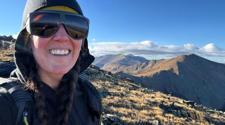 Woman wearing Ibex Mammoth Full-Zip Hoodie on a mountain ridge, smiling with sunglasses and braided hair.