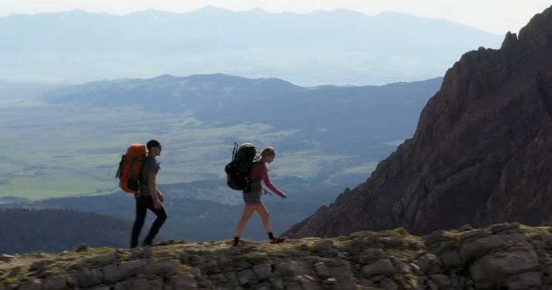 Man and woman hiking along a rocky ridgeline with Mystery Ranch Bridger 55 backpacks, mountain range in the distance.