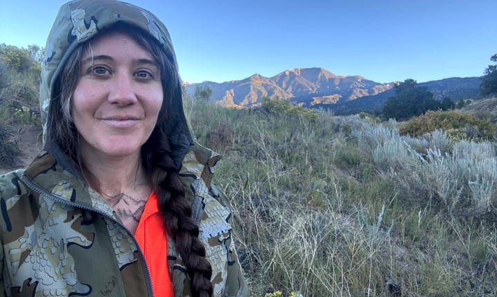 Person wearing KUIU Kenai Hooded Jacket in camouflage while hiking through mountain terrain at sunrise.
