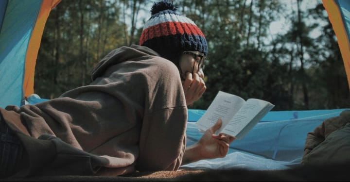 Person lying inside a camping tent reading a book while wearing a knit beanie, with a forest visible through the tent opening.