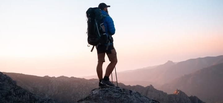 Solo backpacker standing on a rocky summit at sunrise, looking out over distant mountain ranges with trekking poles.